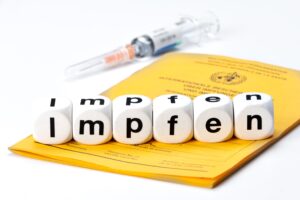 Spritze und Impfausweis. Würfel mit Buchstabenaufdruck aneinandergereiht zeigen das Wort "Impfen"