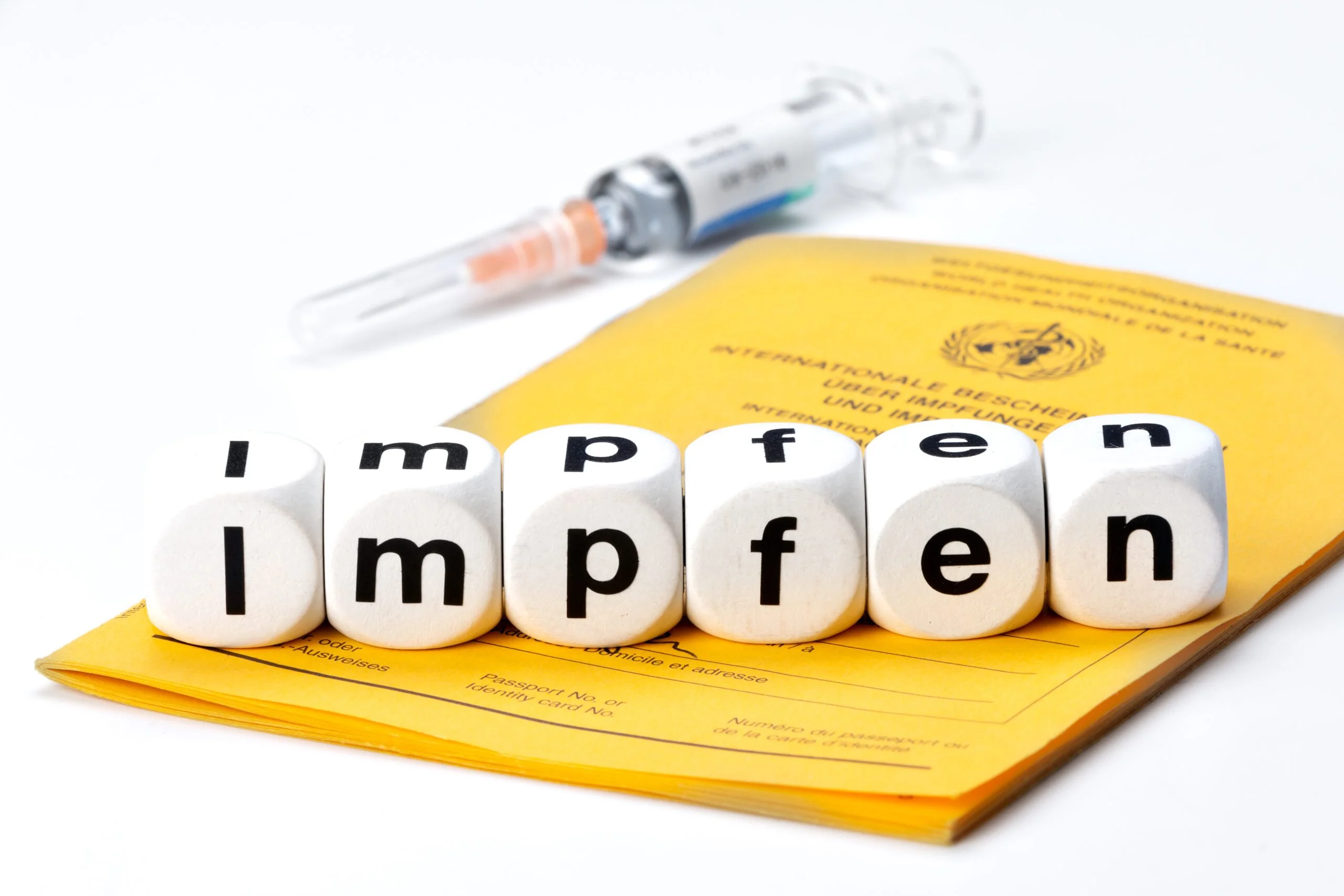Spritze und Impfausweis. Würfel mit Buchstabenaufdruck aneinandergereiht zeigen das Wort "Impfen"